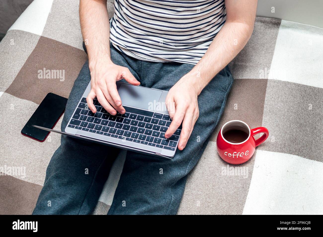 Young man working on a laptop in bed at home and big red cup beside.A man looks at a smartphone. Concept Working remotely Stock Photo