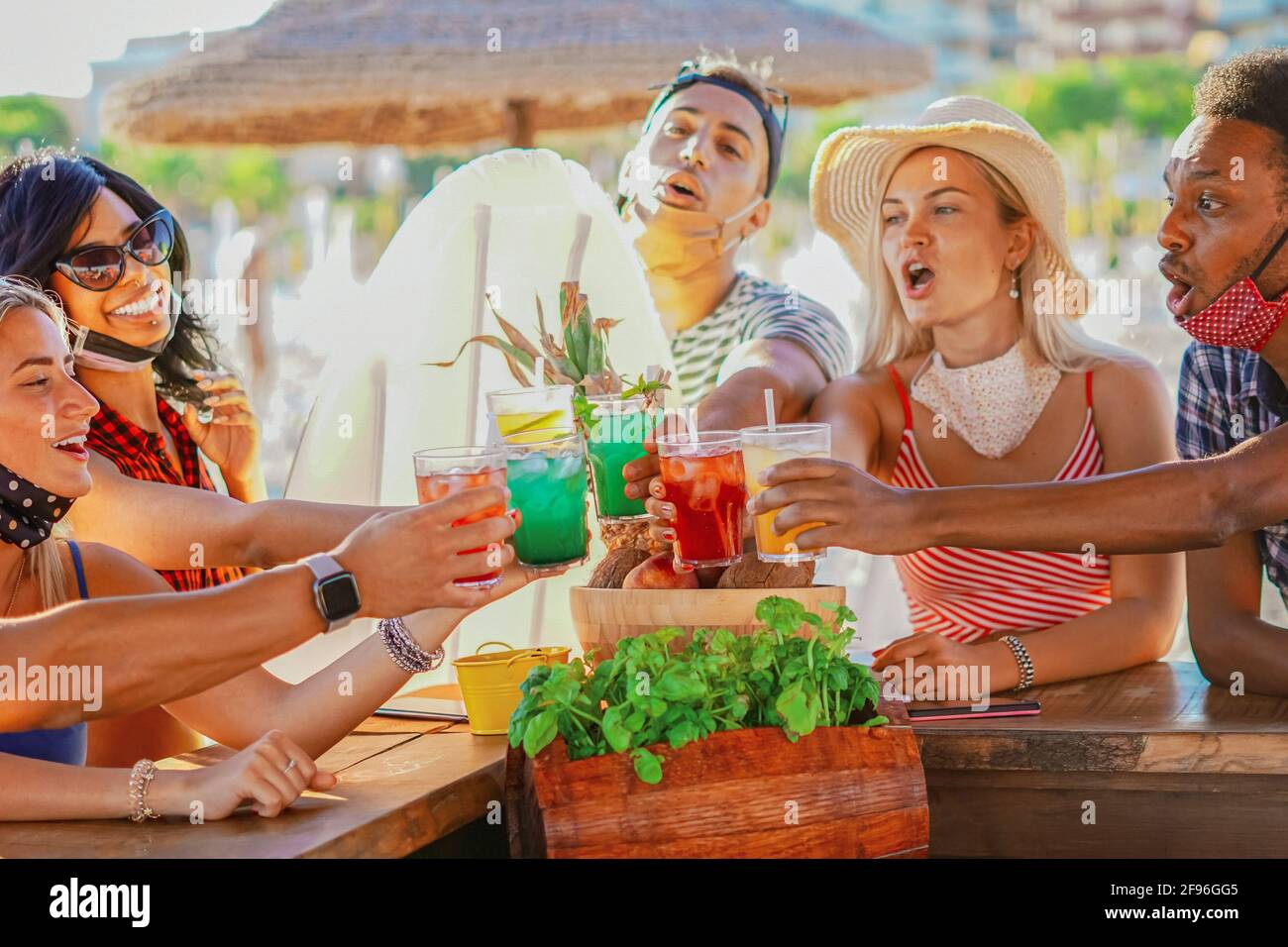 Multiethnic friends drinking coktails in a beach bar outside in summer days with face mask on to be protected from coronavirus -  Happy people cheerin Stock Photo