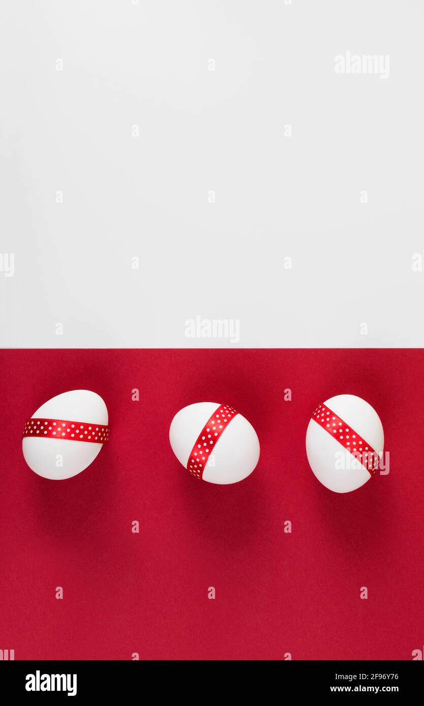 Easter Concept. White Easter eggs decorated with red ribbon on red and white paper background. Vertical photo. Copy space. Flat lay. Stock Photo