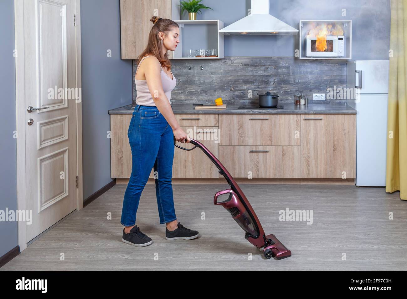 Microwave oven burned out as result of misuse, young woman vacuums floor and does not notice that house fire has started, household appliances have Stock Photo