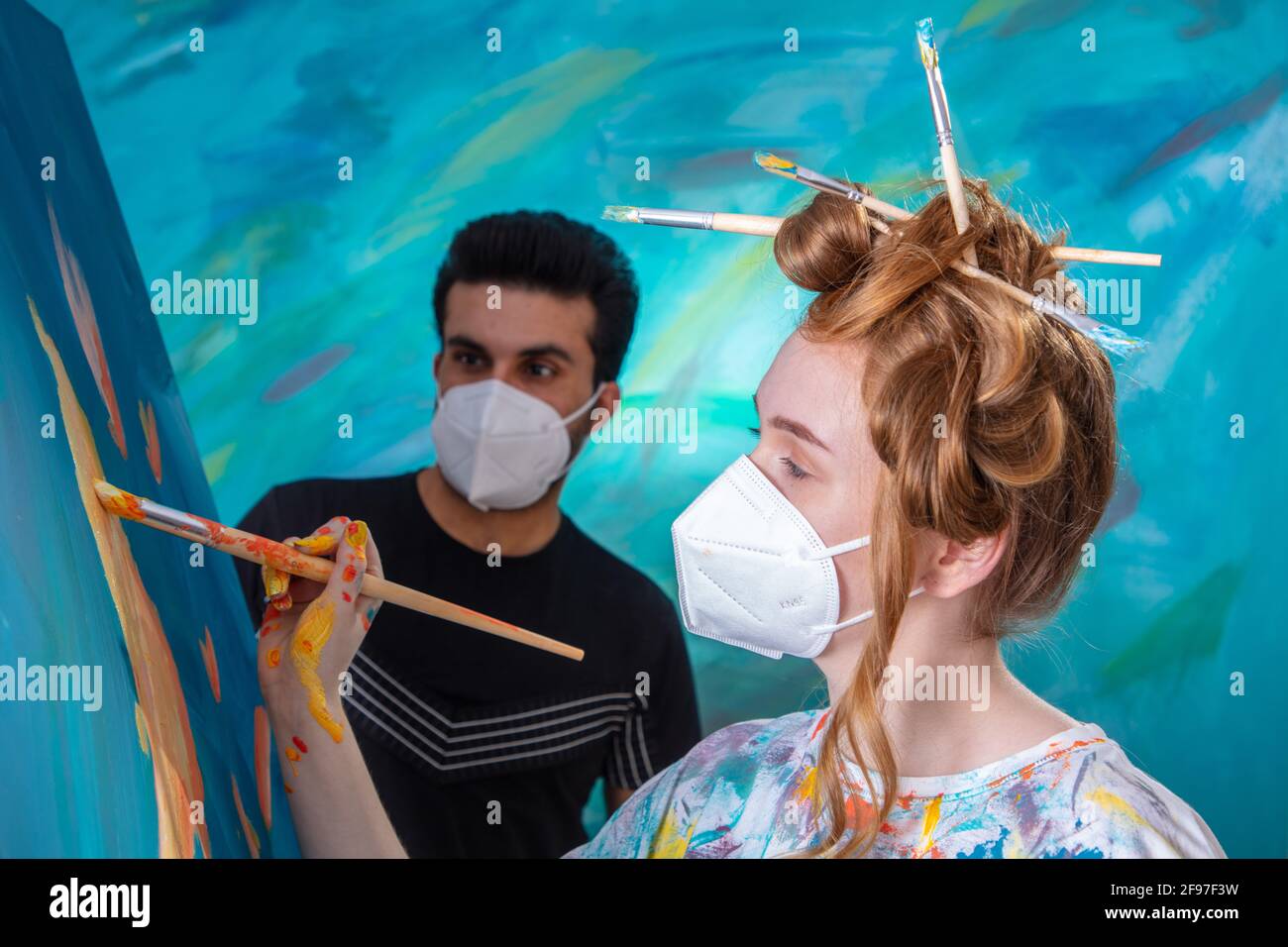 Young woman with updo and brush in her hair while painting, man is watching her, both are wearing FFP2 masks Stock Photo