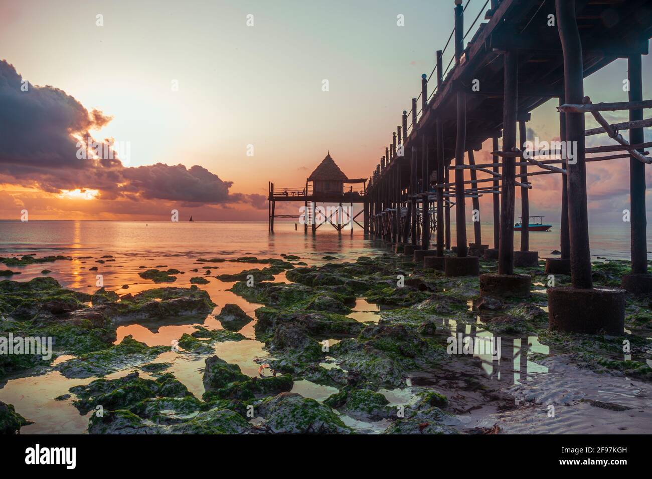 Beautiful Sunrise and a wooden bridge or jetty leading to a Restaurant / Bar called Sansibar at Jambiani beach, Zanzibar, Tanzania, Africa at low tide with green rocks visible in the water Stock Photo