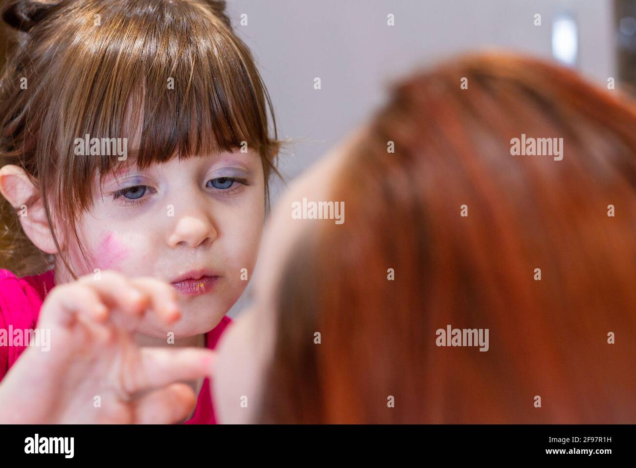 Portrait of a cute, blue-eyed, brown-haired girl with makeup in her face playing to put makeup in a redhead woman Stock Photo