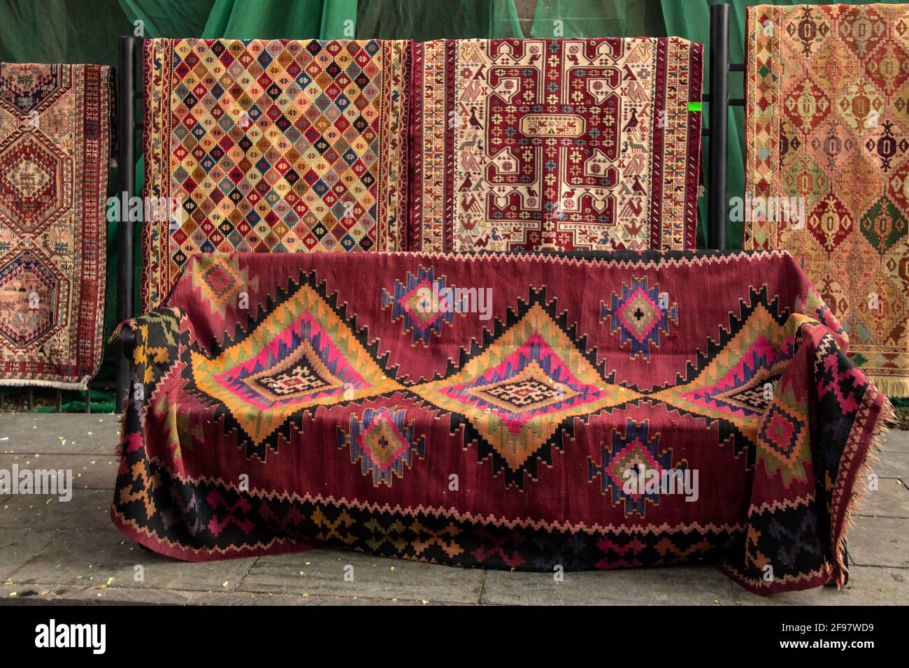 Turkish carpets displayed on the street some hanging and one draped over a seat with flower petals from the trees fallen around Stock Photo