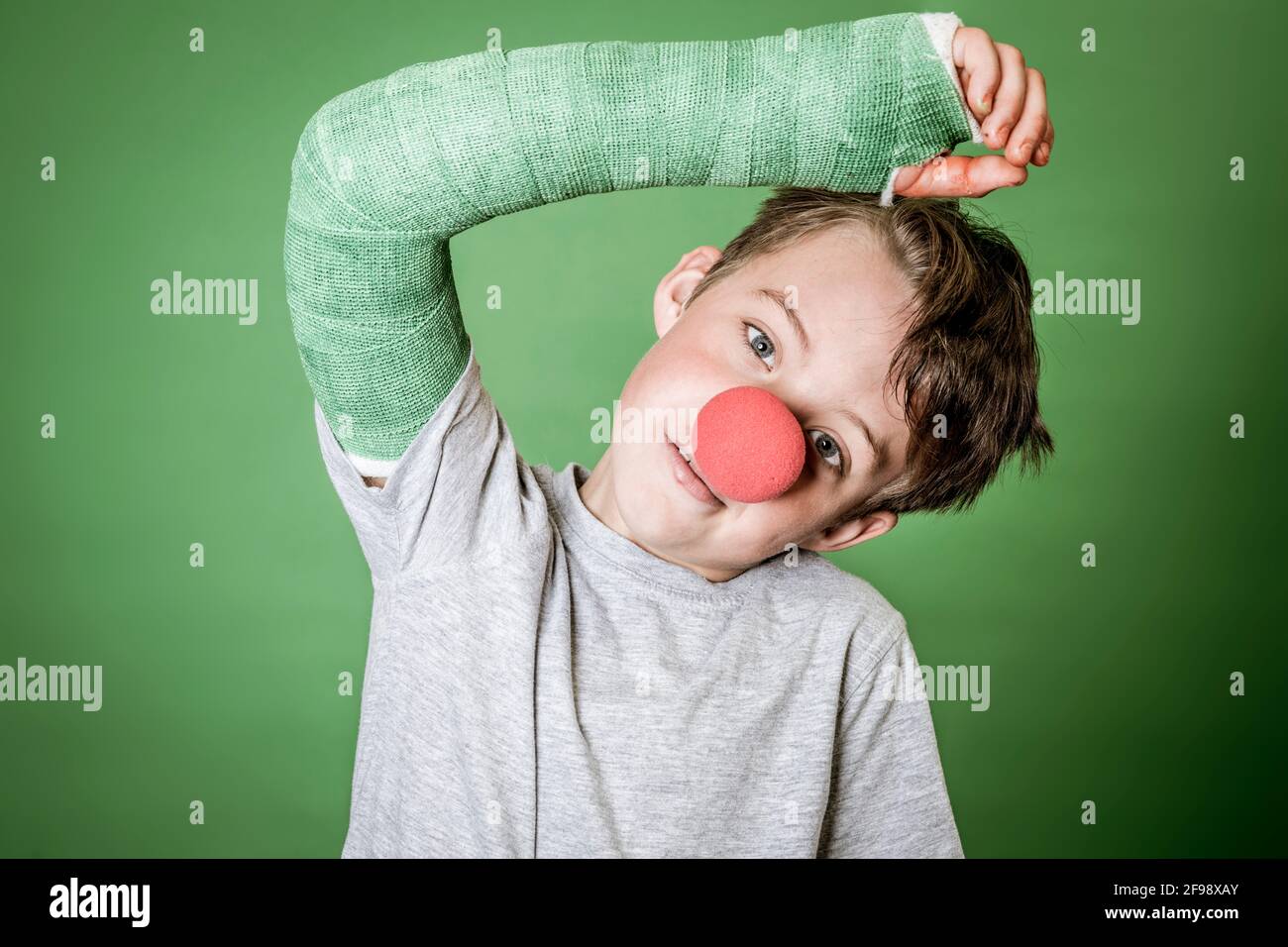 cool, cute schoolboy with green hand plaster and red clown nose is posing in front of green background Stock Photo