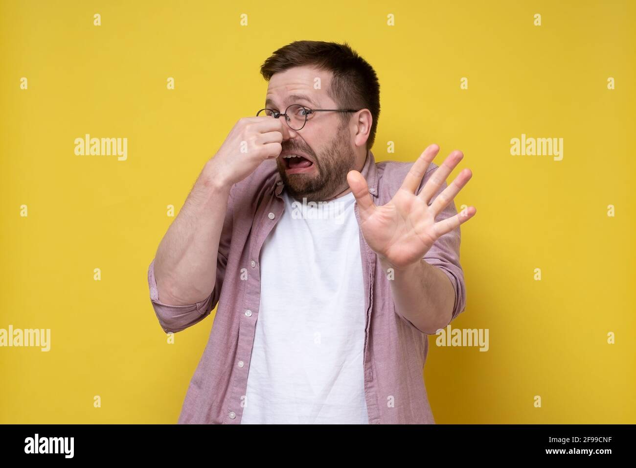 Young man in glasses squeezes nose with fingers because of an unpleasant smell, makes a stop gesture and looks frightened.  Stock Photo