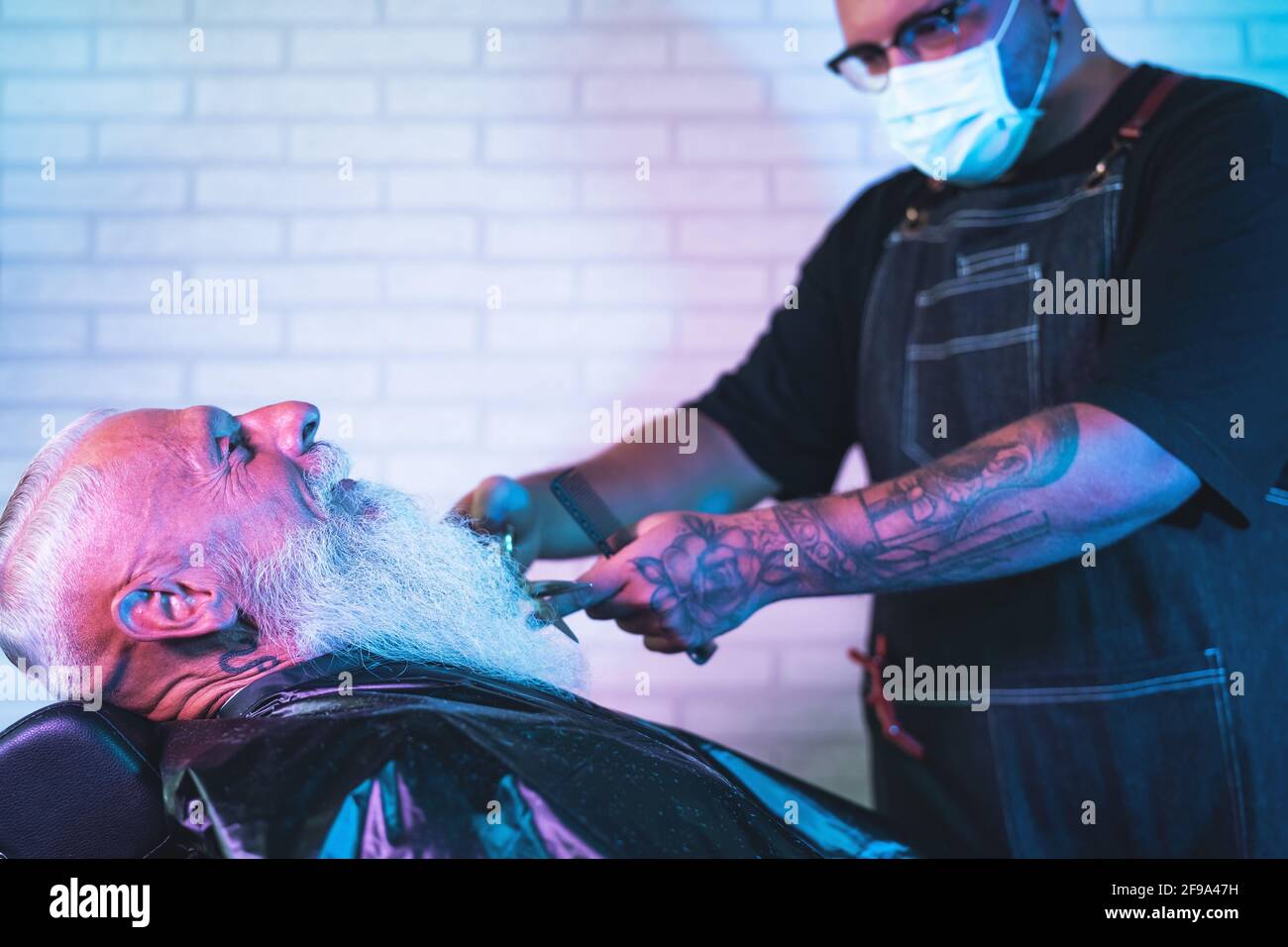 Male hairdresser cutting beard to hipster senior client - Young hairstylist working in barbershop - Haircut profession concept Stock Photo