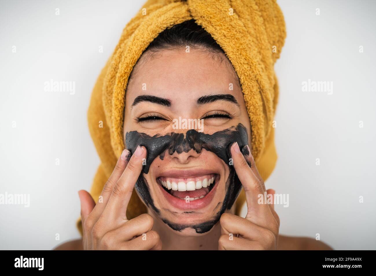 Young smiling woman applying charcoal mask on face - Happy girl having skin care spa day at home Stock Photo