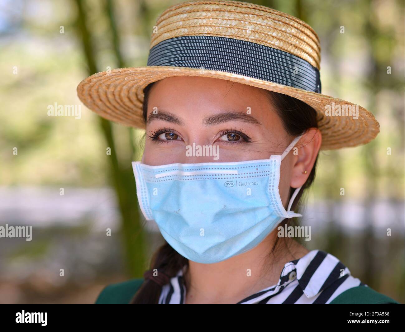 Beautiful young Mexican woman wears a stylish Panama straw hat and poses with a light-blue surgical face mask during the global coronavirus pandemic. Stock Photo