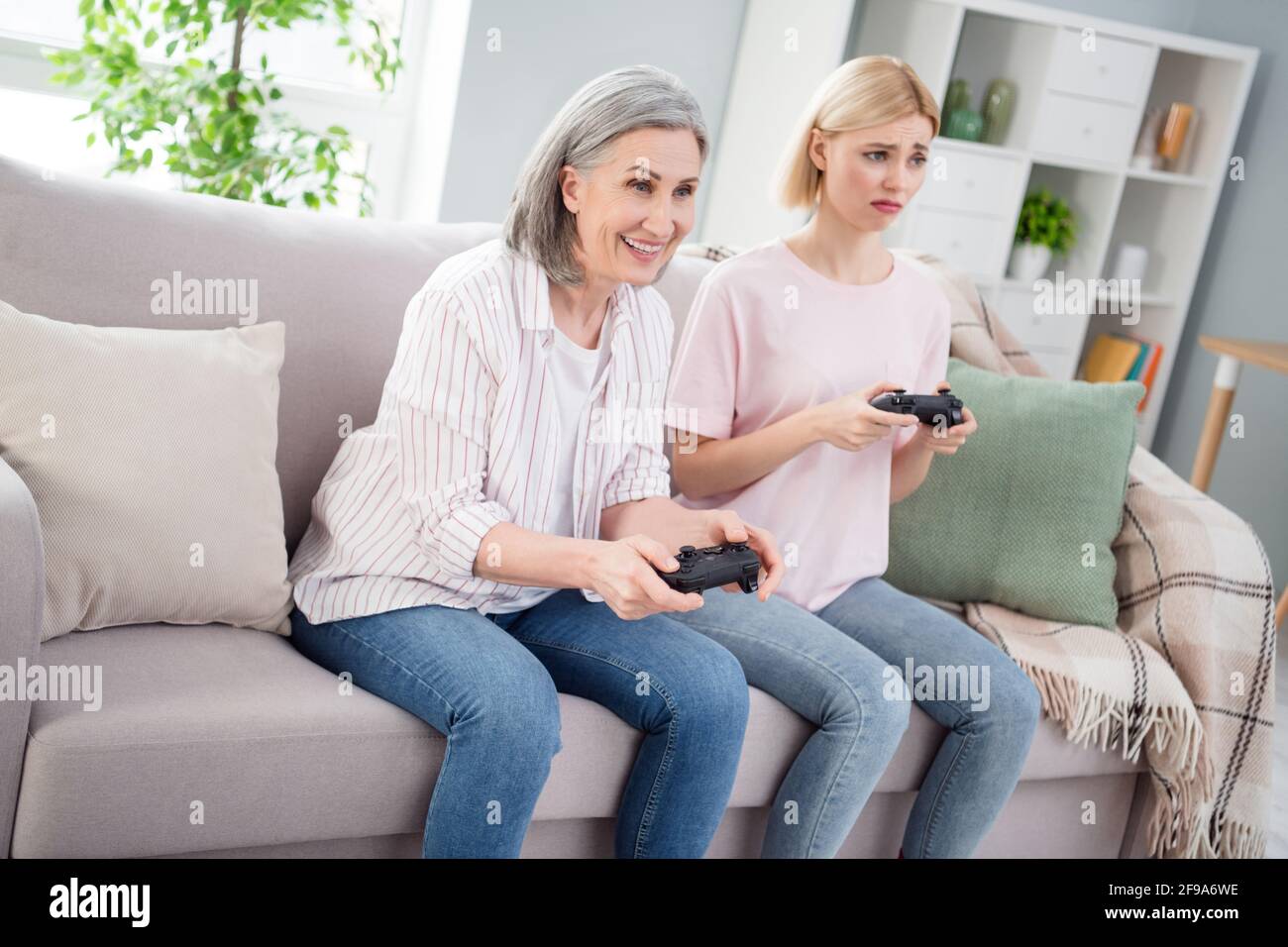 Photo of happy grandmother and upset young woman sit sofa play game joystick weekend indoors inside house Stock Photo