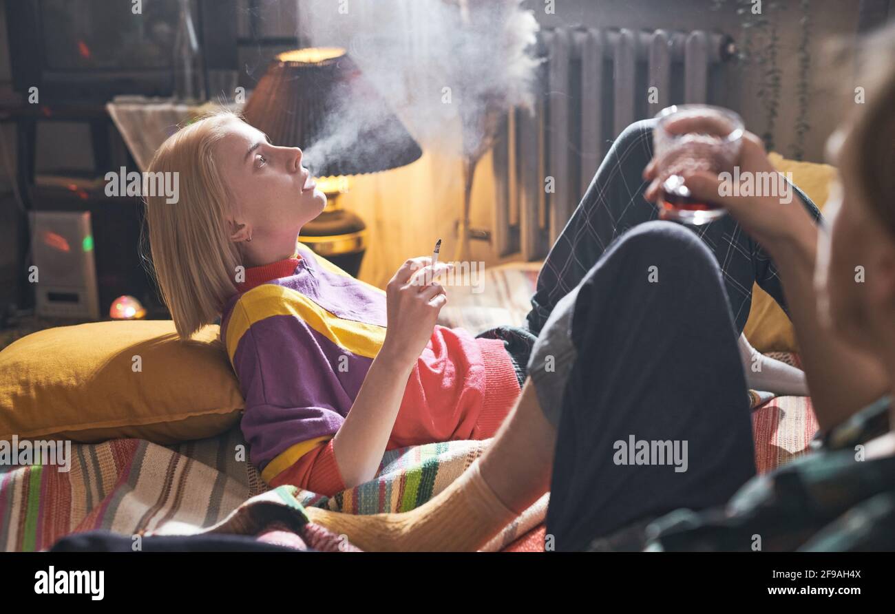 boyfriend smokes cigarettes Young woman lying on bed and smoking a cigarette with her boyfriend sitting  on the bed and drinking wine Stock Photo