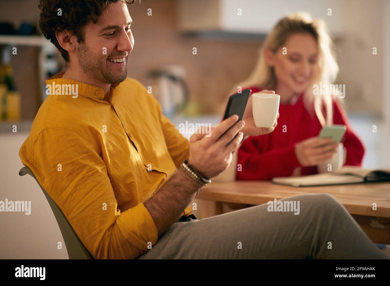 young adult caucasian couple laughing watching their cell phones in the morning, drinking coffee. Modern social distraction concept Stock Photo