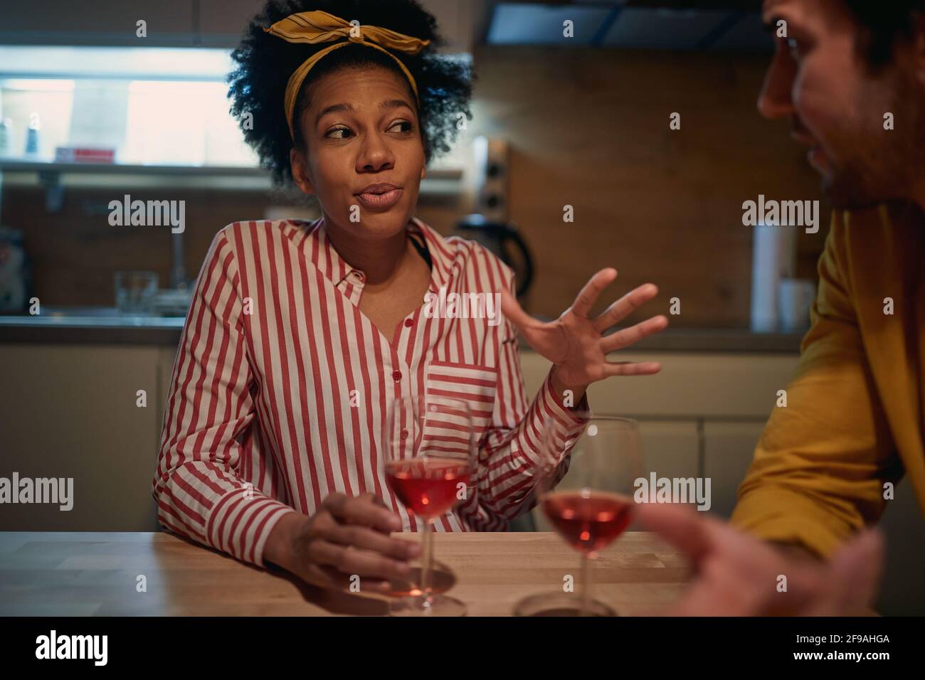 young adult afro-american woman with scarf on her head, talking with caucasian man, explaining, drinking red win. Friendship at home during coronaviru Stock Photo