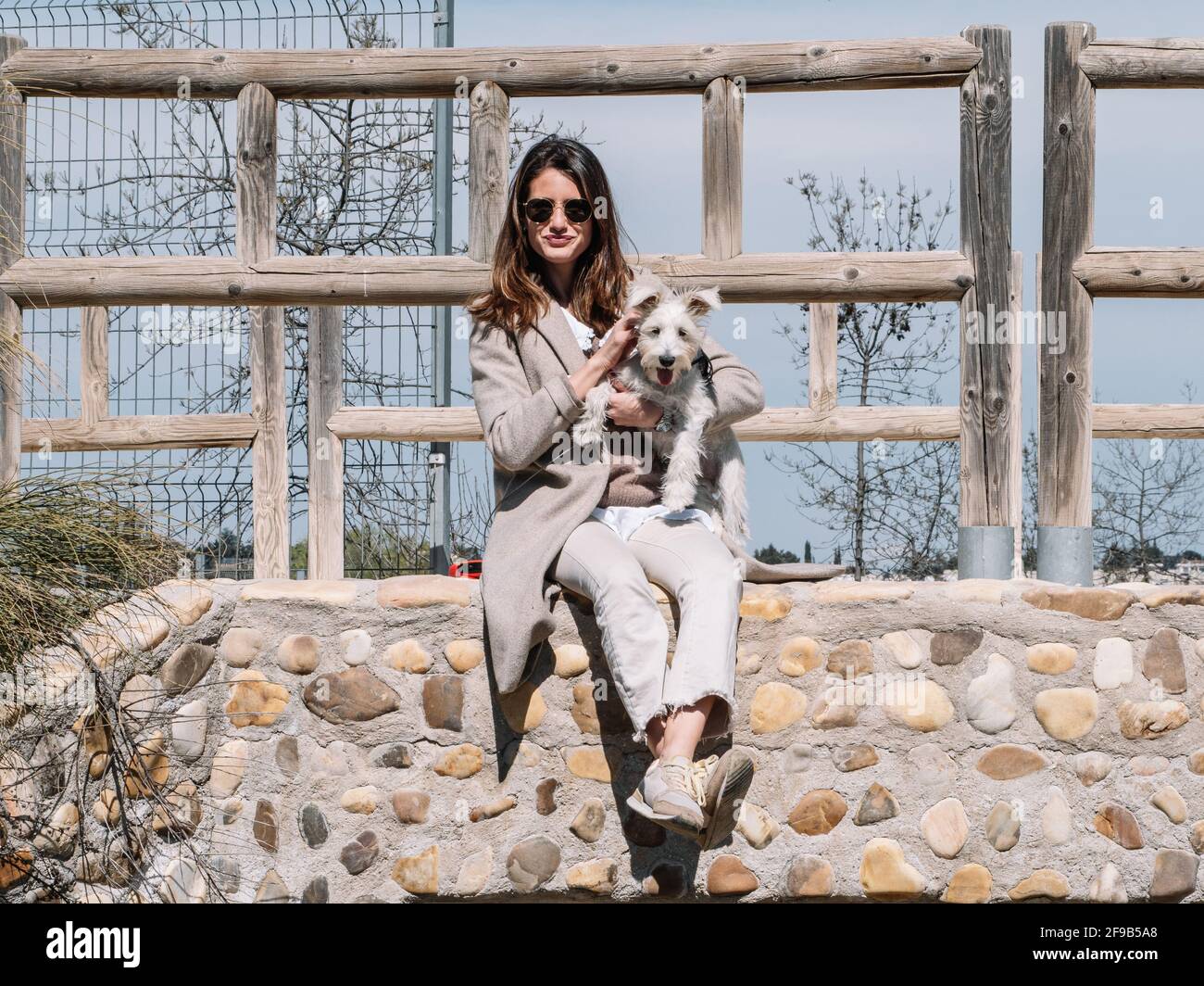 Young woman sitting on a stone fence with her Schauzer dog, poses and touches her hair looking straight ahead. Stock Photo