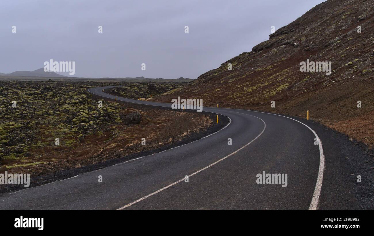 Diminishing perspective of empty winding country road (s-shape) beside moss covered lava field of volcanic stones near Grindavik, Reykjanes, Iceland. Stock Photo