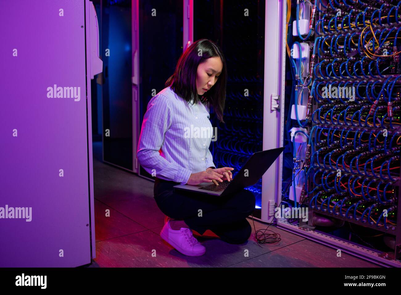 Serious young Asian data center engineer crouching by server equipment and using laptop while troubleshooting problem Stock Photo