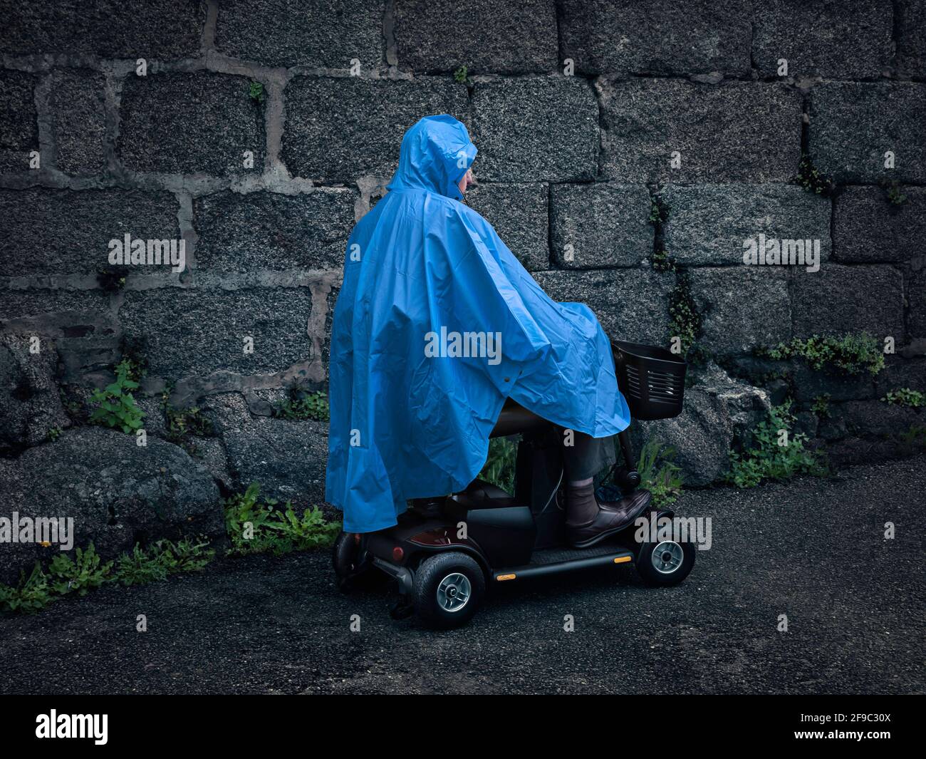 Man on a Mobility Scooter / Vehicle wearing a Blue Raincoat / Poncho / Cagoule / Jacket, in profile with Old Grey Stone Wall Background Stock Photo