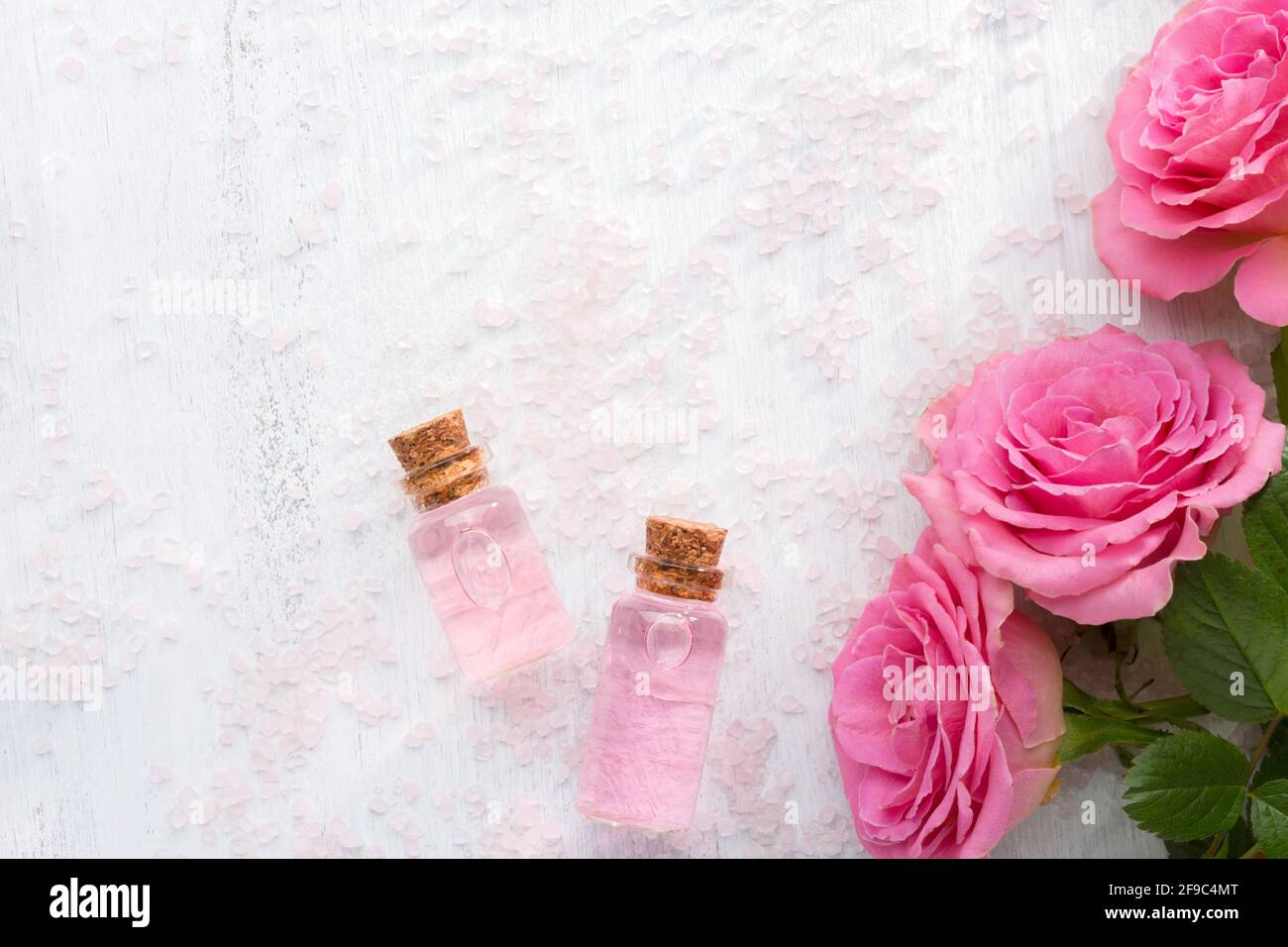 Two bottles with rose oil, crystals of mineral bath salts and pink roses on the white  wooden table. Stock Photo