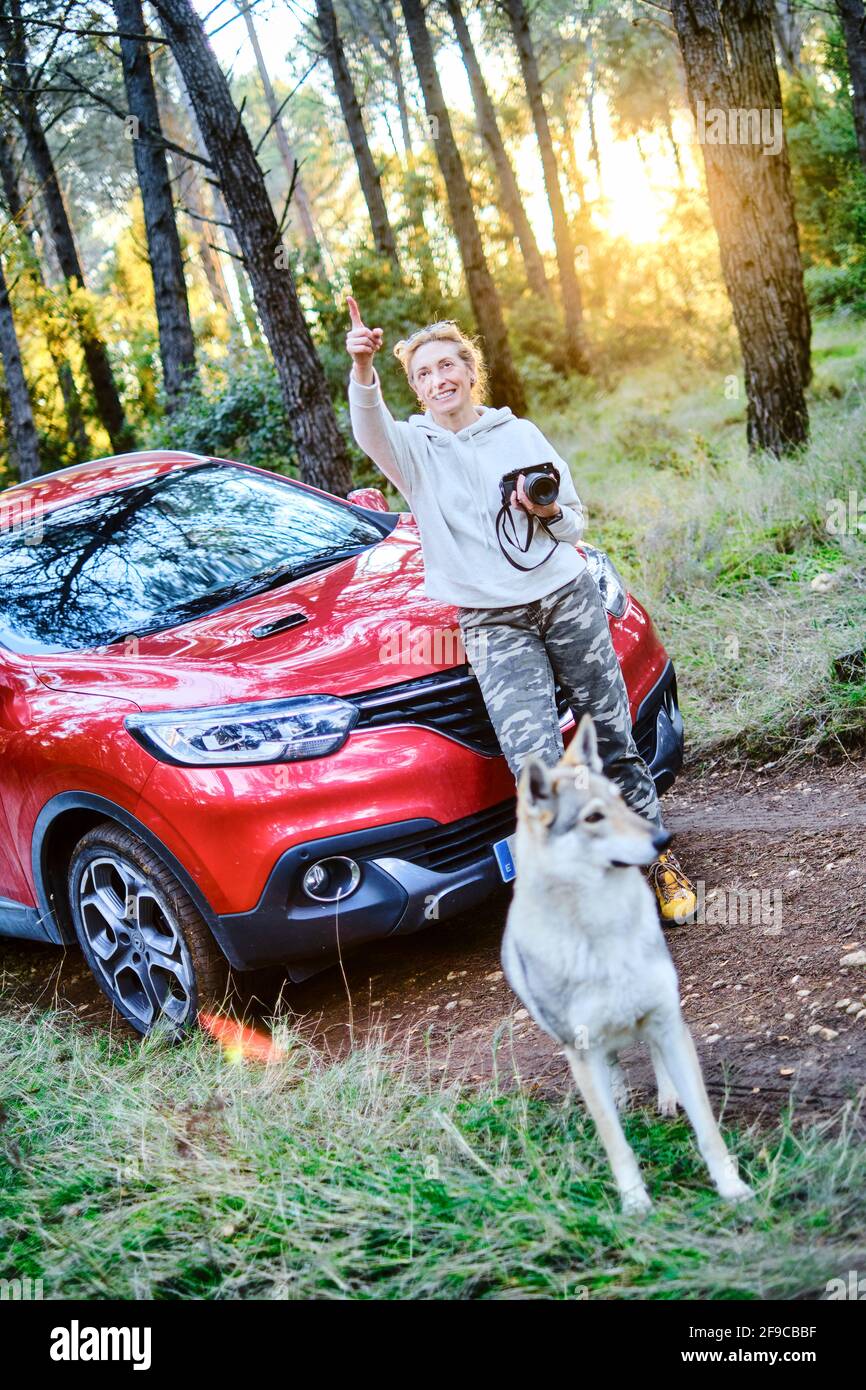 Young adult caucasian woman taking pictures at sunset with a dog and close to a car parked in a forestry path in autumn. Travels concept. Stock Photo