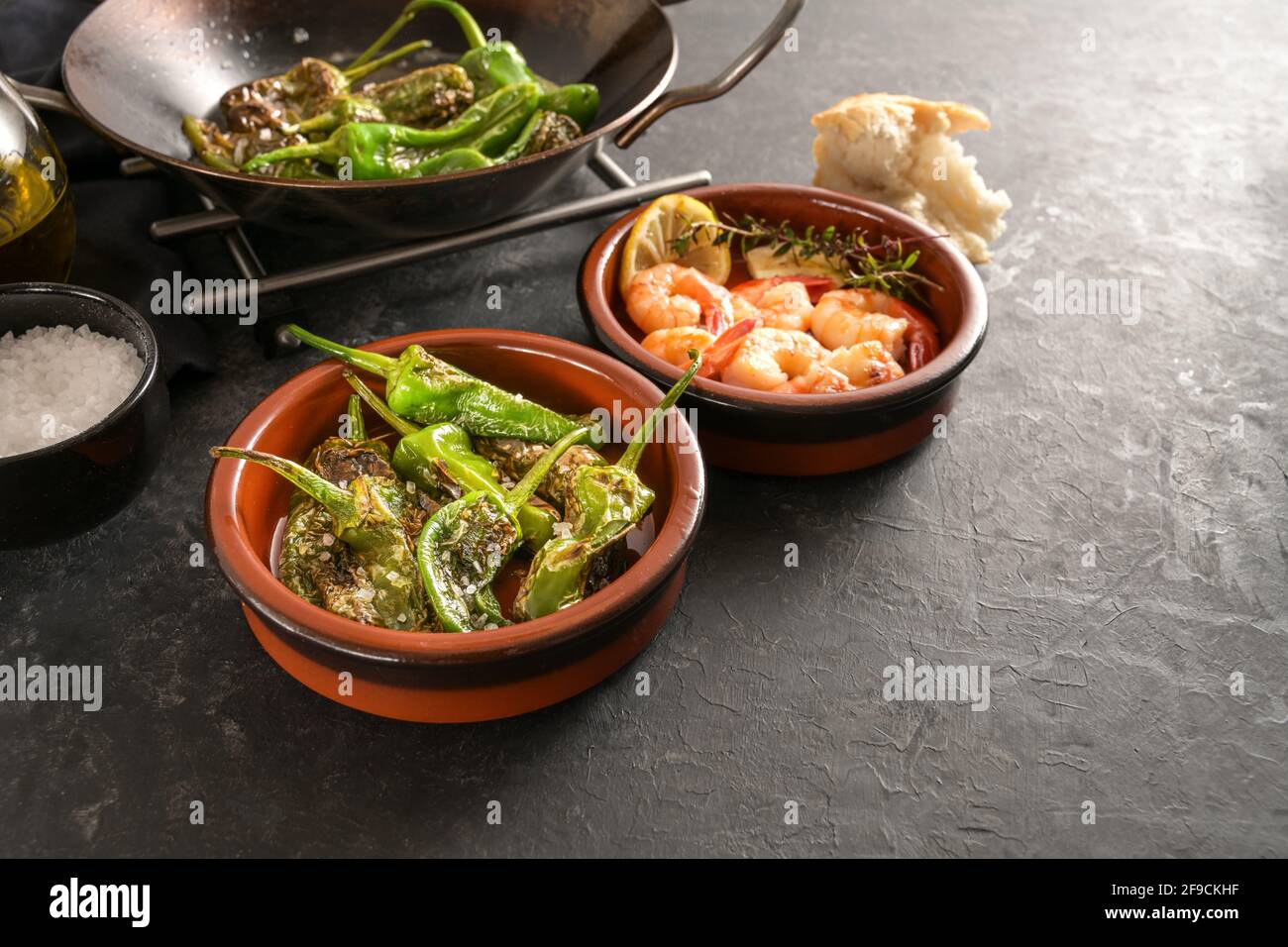 Spanish tapas, fried pimientos or padron peppers and shrimps in traditional bowls on a dark gray slate background, copy space, selected focus, narrow Stock Photo