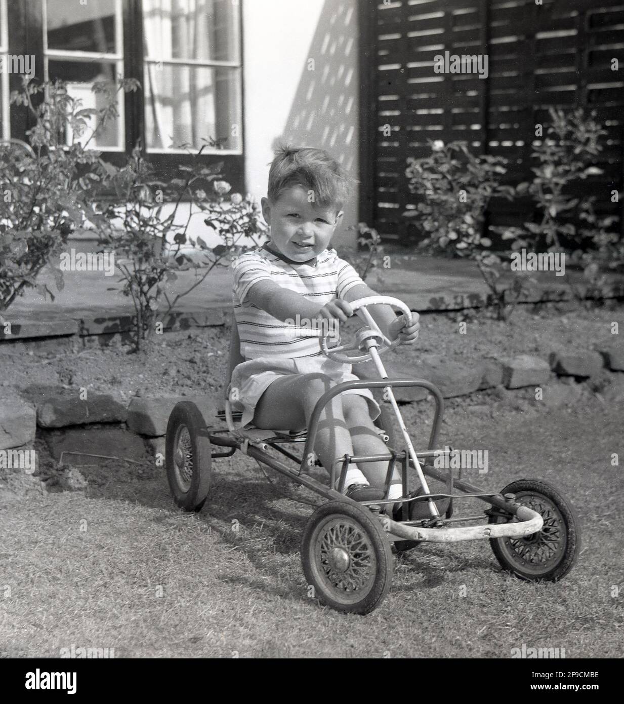 1966, historical, outside in a garden, a young boy, sitting, with his hands on the plastic steering wheel, in a four-wheeled metal-framed pedal toy car. As it is minus the outer shell - maybe broken over time - the toy could be a hand-med-down. Or looking at it, quite possibly a DIY home-made machine made from spare pieces of metal and four old pram wheels. Stock Photo