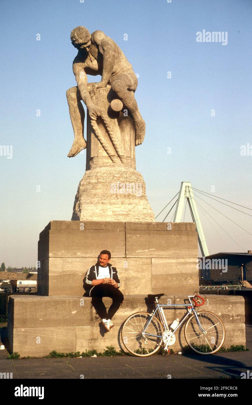 Cyclist resting beside the Tauzieher (Rope Puller) sculpture beside the Rhine, Cologne, North Rhine-Westphalia, Germany Stock Photo