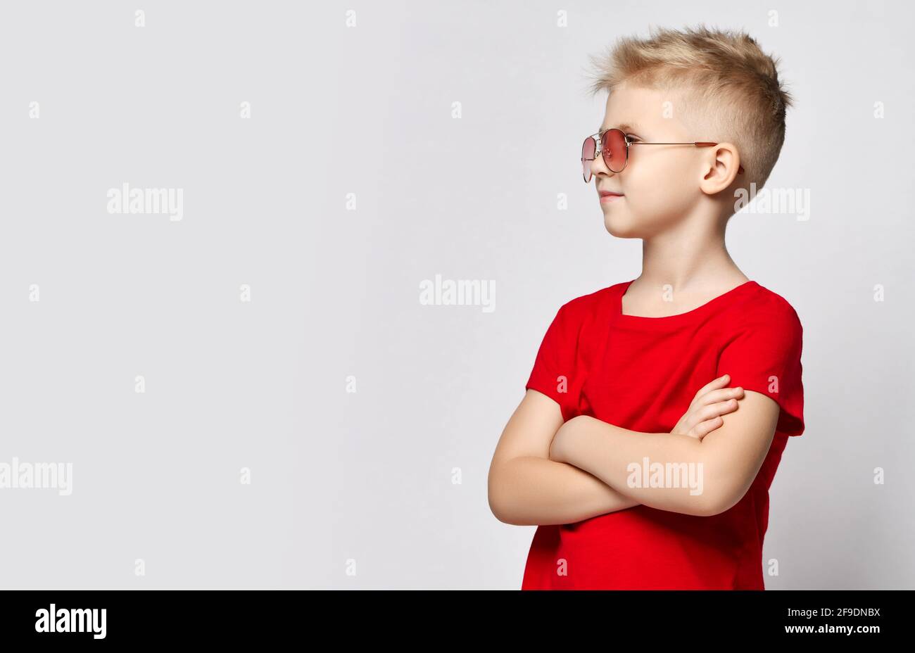 Portrait of blonde kid boy in red t-shirt and sunglasses standing with arms crossed at chest, looking at copy space Stock Photo