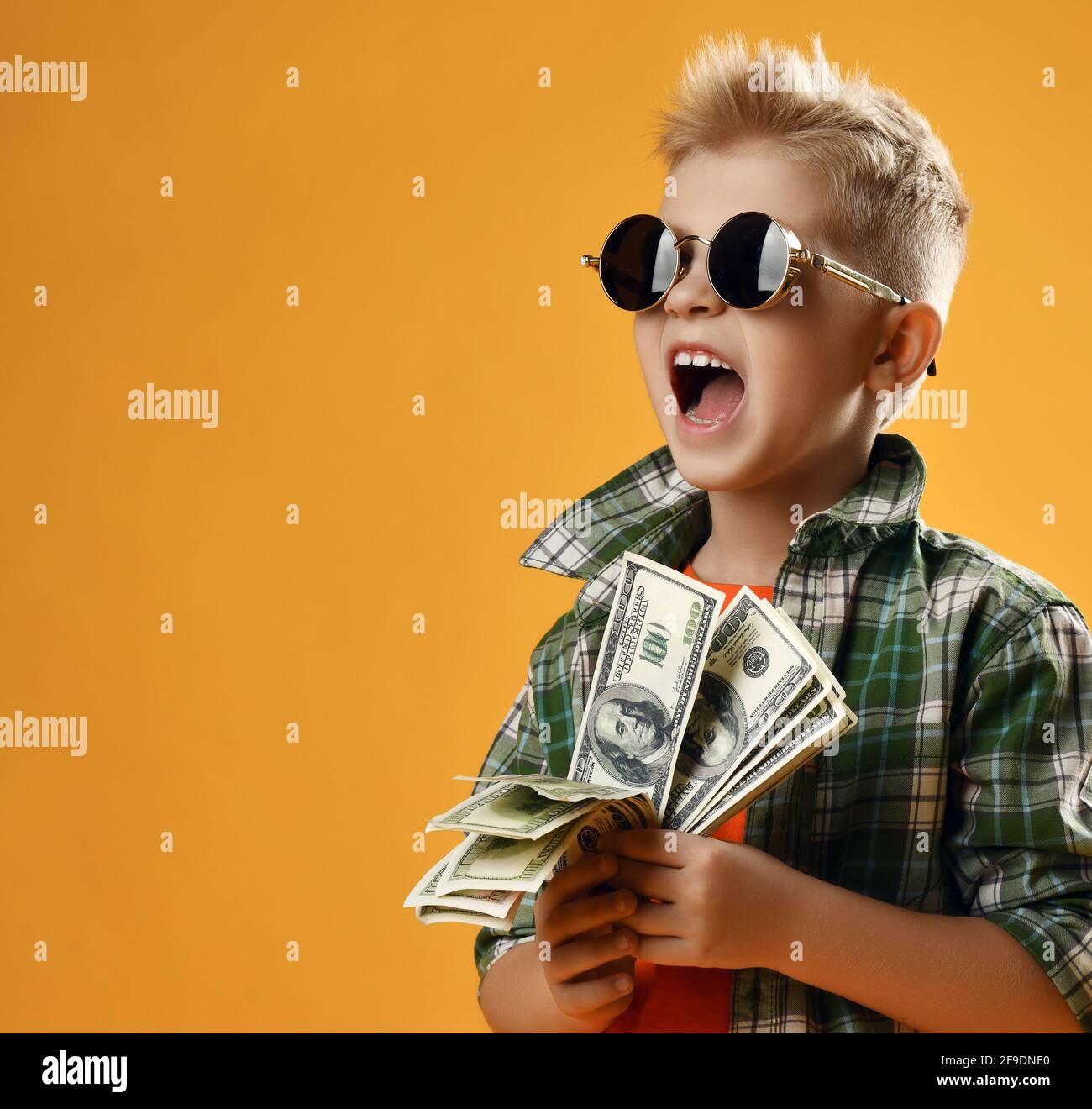 Blonde rich kid boy in round sunglasses and checkered plaid shirt stands with fan of dollar cash in hands and screams Stock Photo