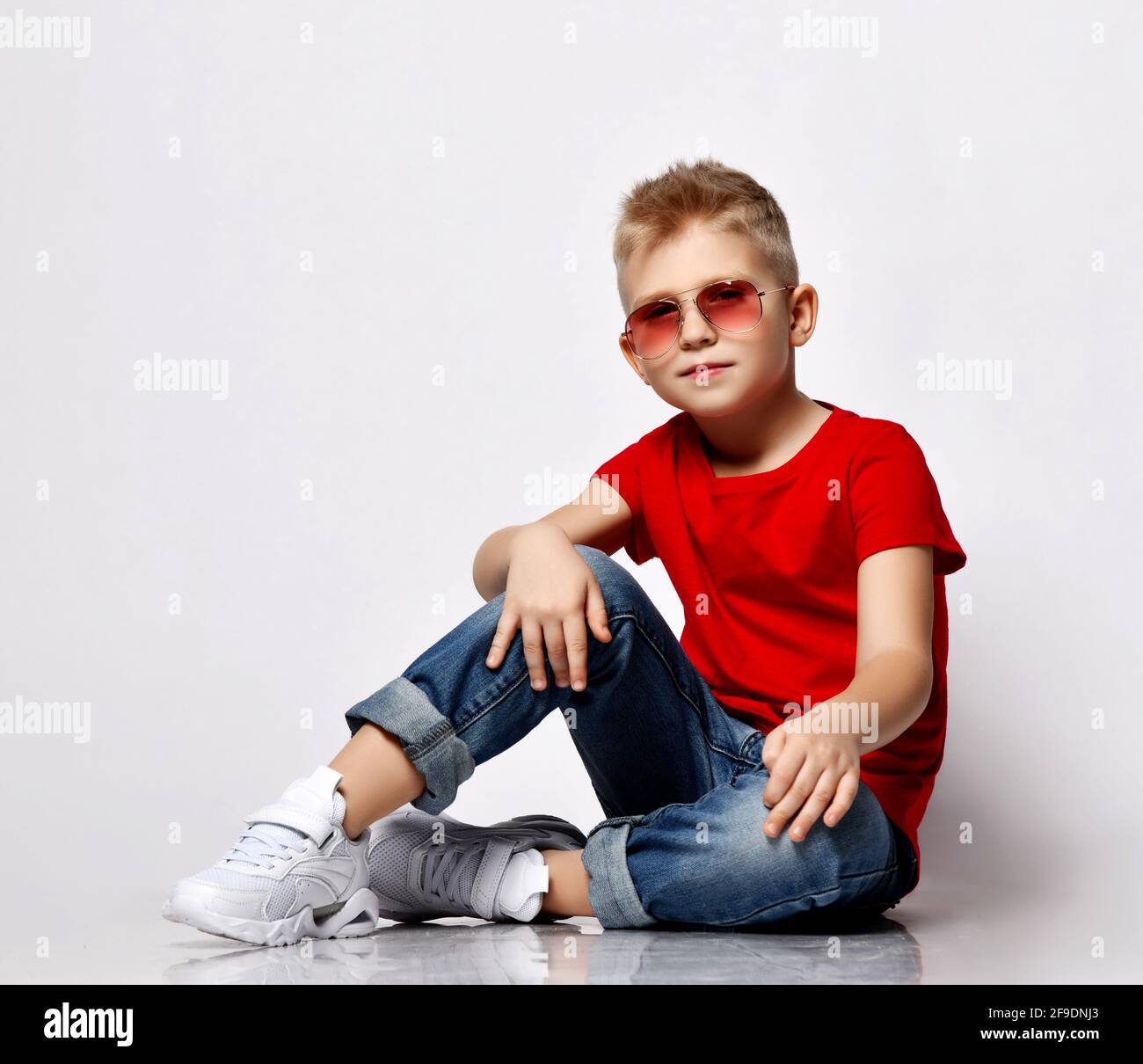 Cool blonde kid boy leader in red t-shirt, blue jeans, white sneakers and sunglasses sits on floor in relaxed pose Stock Photo
