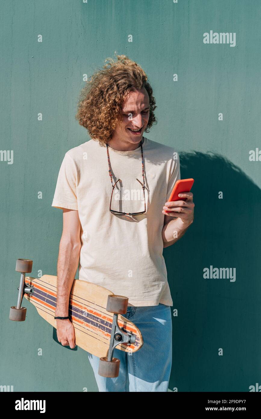 vertical portrait of a long haired young man checking his phone. He wears casual clothes and holds an skateboard Stock Photo