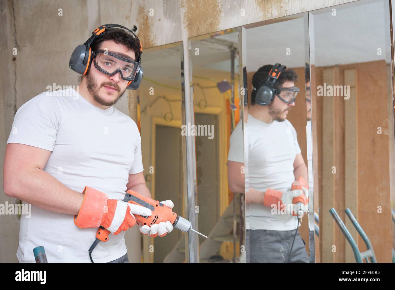 Builder holding a drill and looking at camera at the construction site. Construction worker wearing safety gloves, glasses and earmuffs. Stock Photo