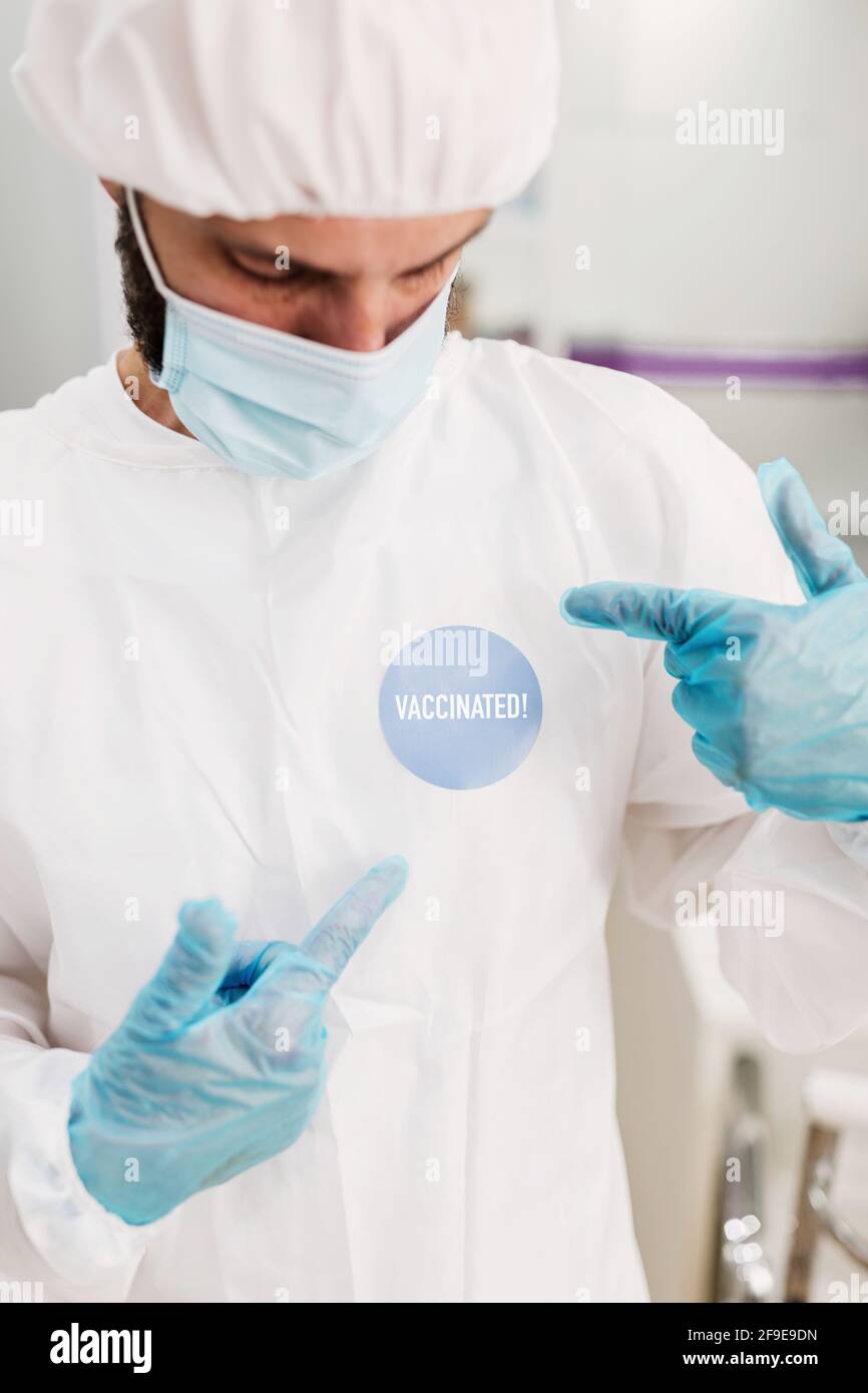 Positive male doctor with latex gloves and protective medical mask pointing at I'm vaccinated message sticker on white uniform standing in modern medi Stock Photo