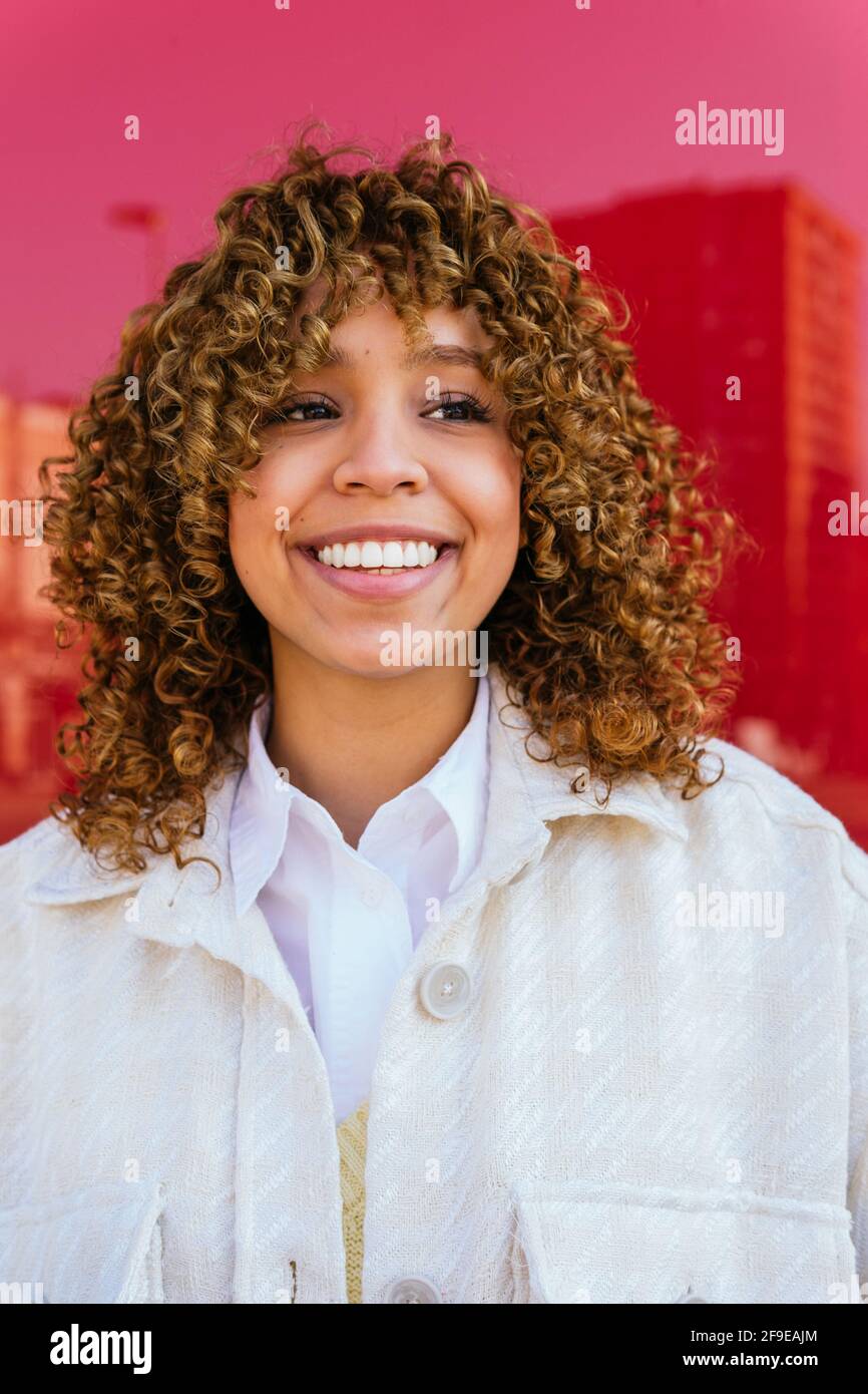 Delighted African American female with curly hairstyle standing on red background in studio looking away Stock Photo
