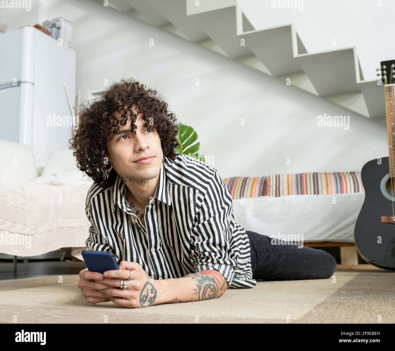 Young contemplative male in striped shirt with tattoos and cellphone lying on carpet while looking up in flat Stock Photo