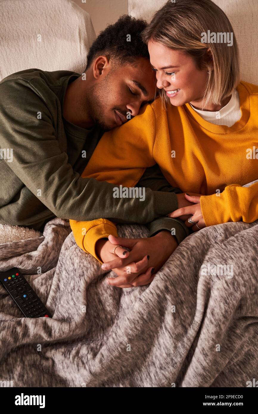 High angle of loving multiethnic couple relaxing on couch under blanket while cuddling and