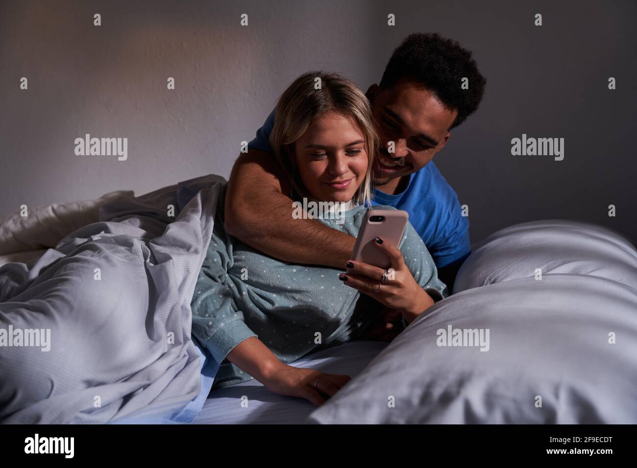 Delighted Multiracial Couple Lying On Bed While Browsing Smartphone And Having Fun Together In