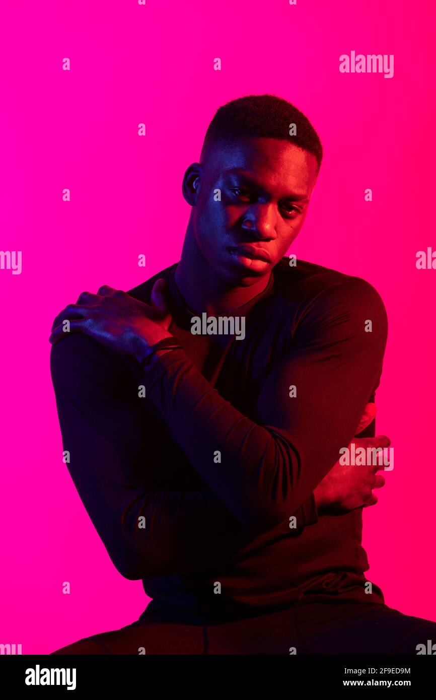 Confident young black man in dark sportive outfit looking at camera on neon pink background in dark studio Stock Photo