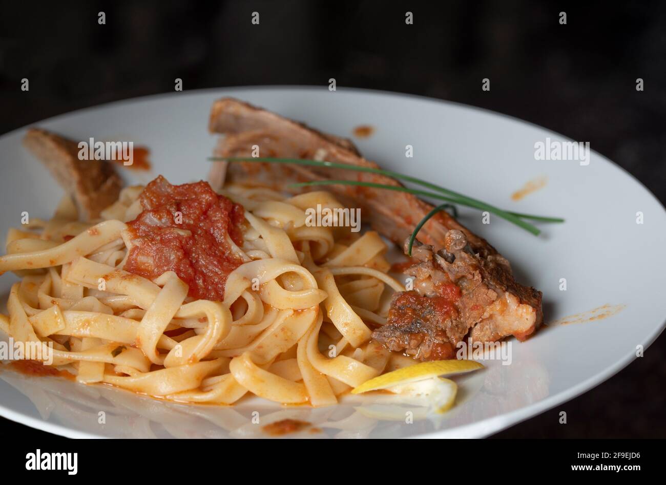 A lunch with lamb ribs on ribbon noodles with tomato sauce in front of a dark background Stock Photo