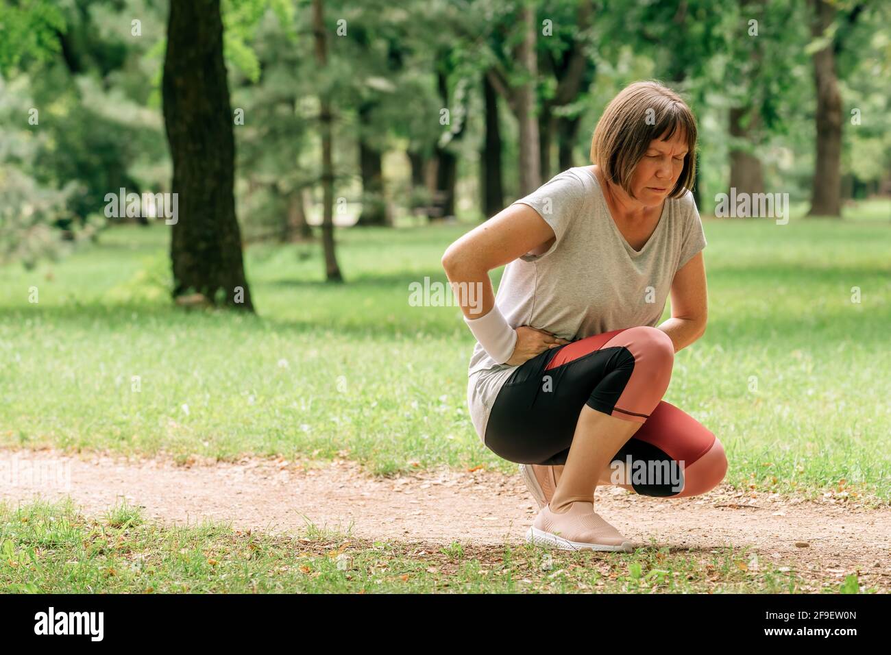 Female jogger with painful face grimace after feeling pain in lower abdomen during running in park Stock Photo