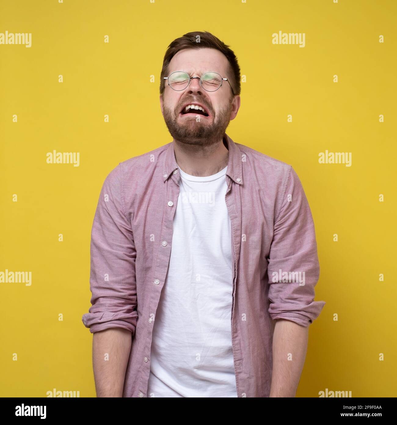 Sobbing, unhappy man in glasses and casual clothes, stands with hands down and eyes closed. Stress concept. Yellow background. Stock Photo