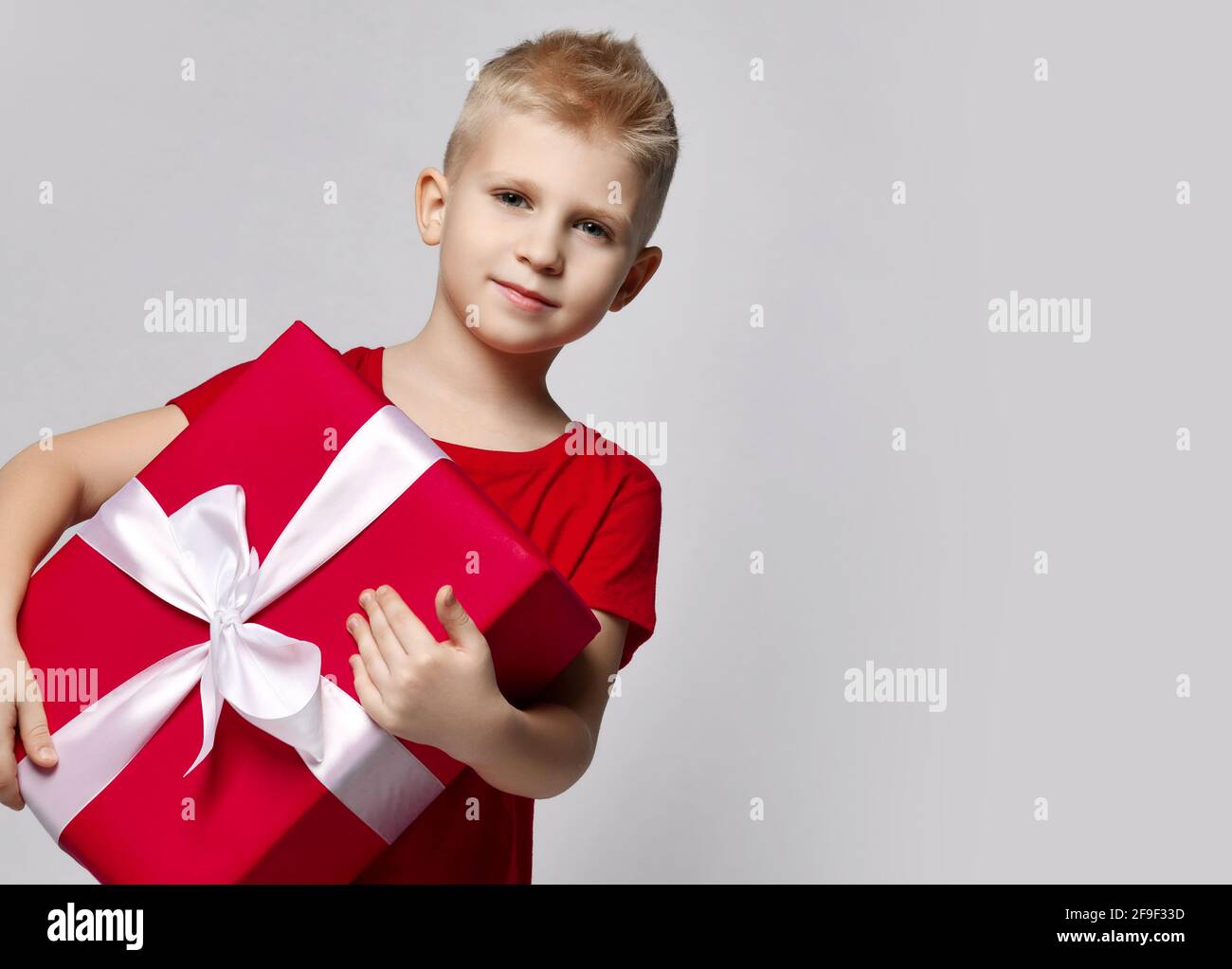 Stylish cheerful boy child in red t-shirt holding big present box with ribbon in hands looking at camera Stock Photo
