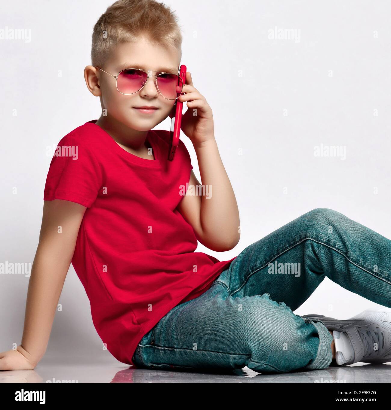 Stylish kid boy child in red t-shirt, jeans, sneakers and sunglasses sitting on floor talking on smartphone Stock Photo