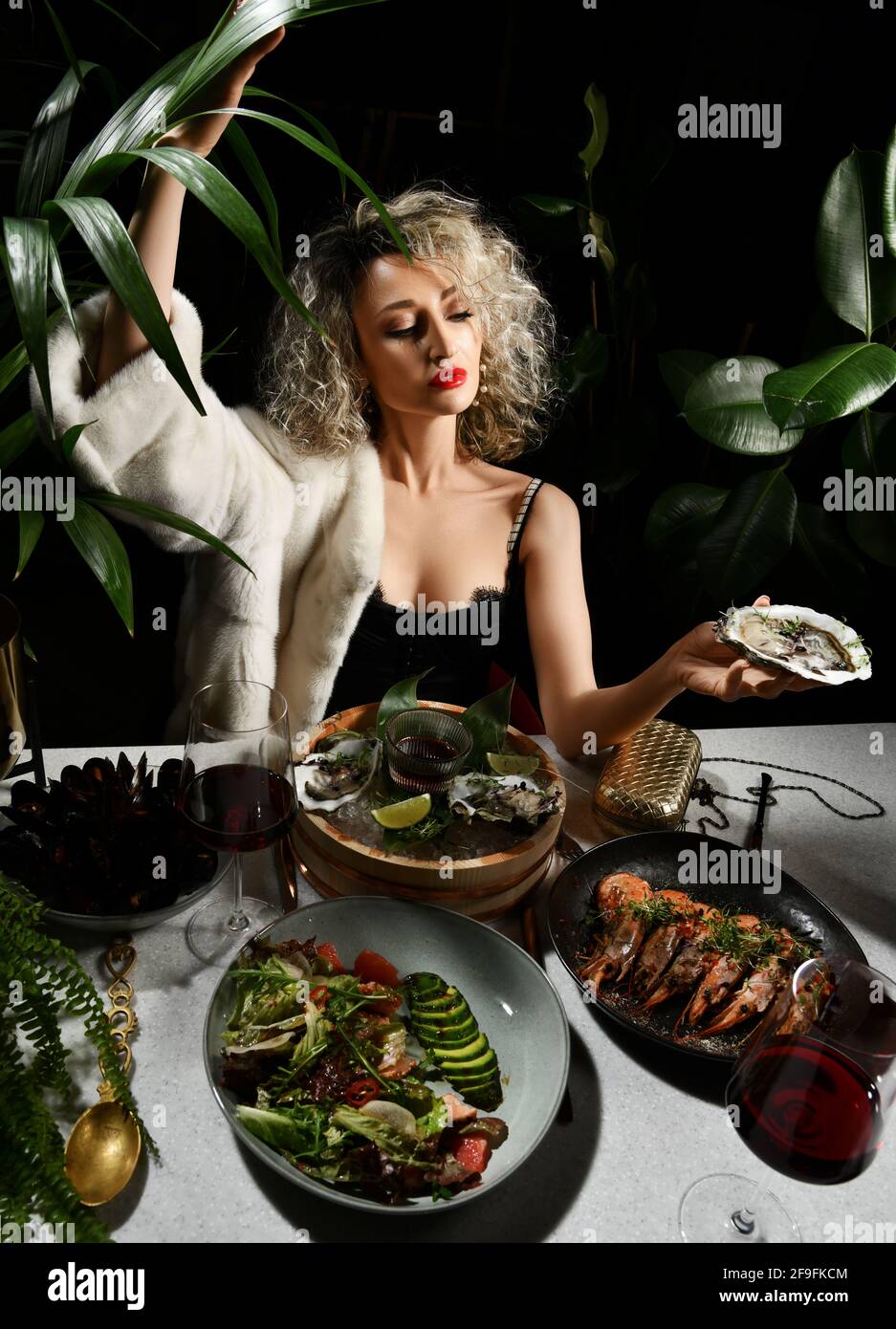 Arrogant picky rich woman in black evening dress is in seafood restaurant sits holding oysters, prawns, mussels in hand Stock Photo