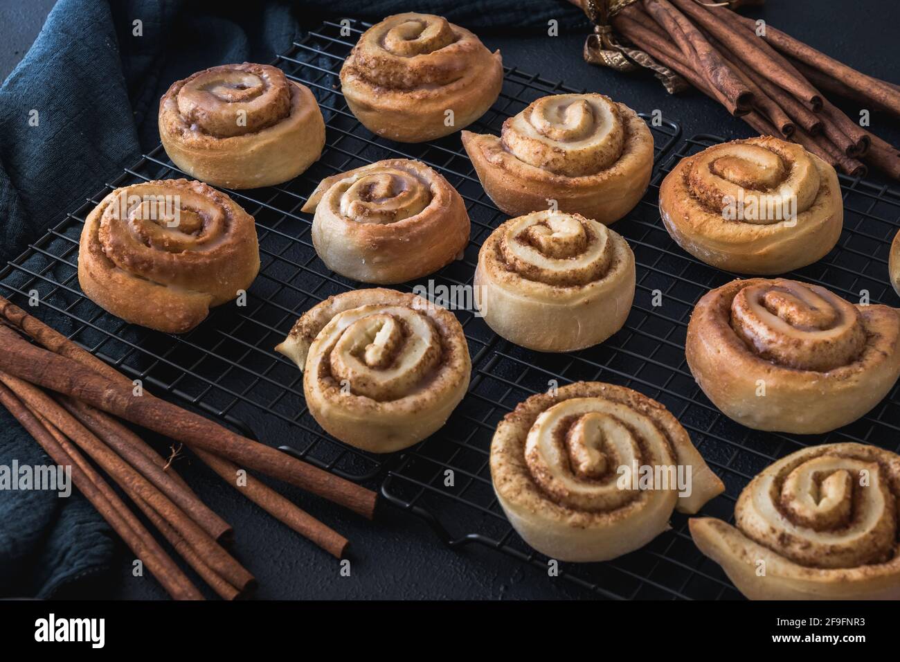 Homemade cinnamon rolls on a cooling rack with cinnamon sticks and dark blue cloth. Stock Photo