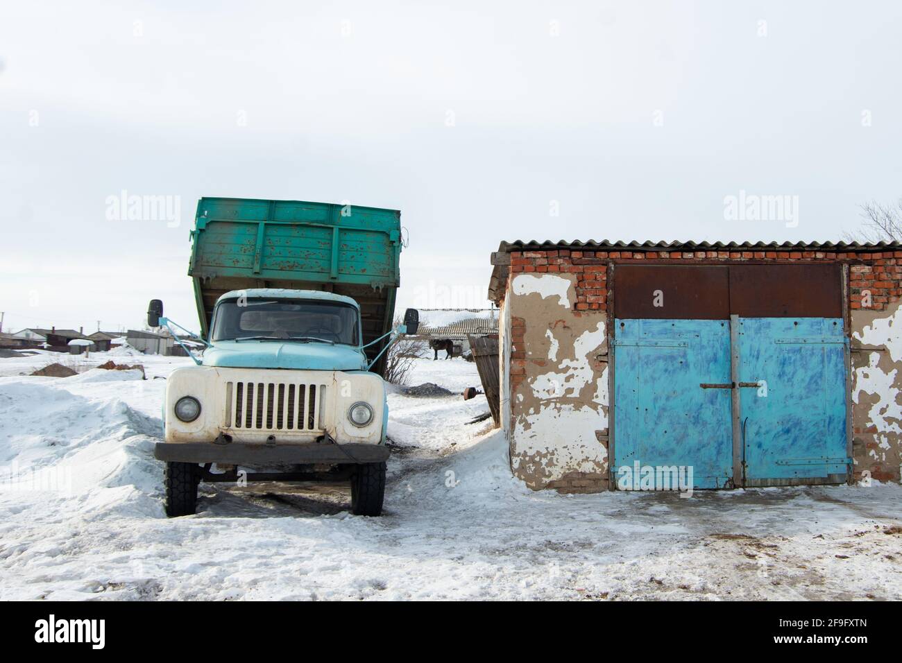 A heavy old blue dump truck is parked next to a building amid white snow, waiting for loading to begin. Delivery of goods in winter Stock Photo