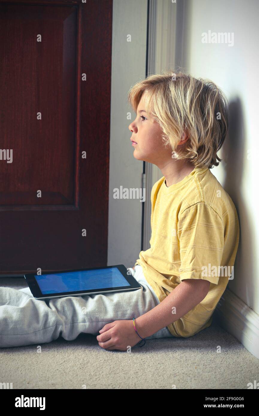 Infant blond boy 4-6 years gazes wistfully out of front door of his home, with smart tablet iPad computer on his lap home missing school and friends Stock Photo