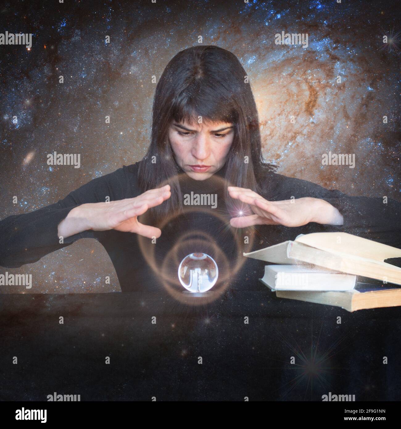 The girl holds her hands over a shining ball and looks to the future next to magic books on the background of galaxie. The concept of paranormal abili Stock Photo