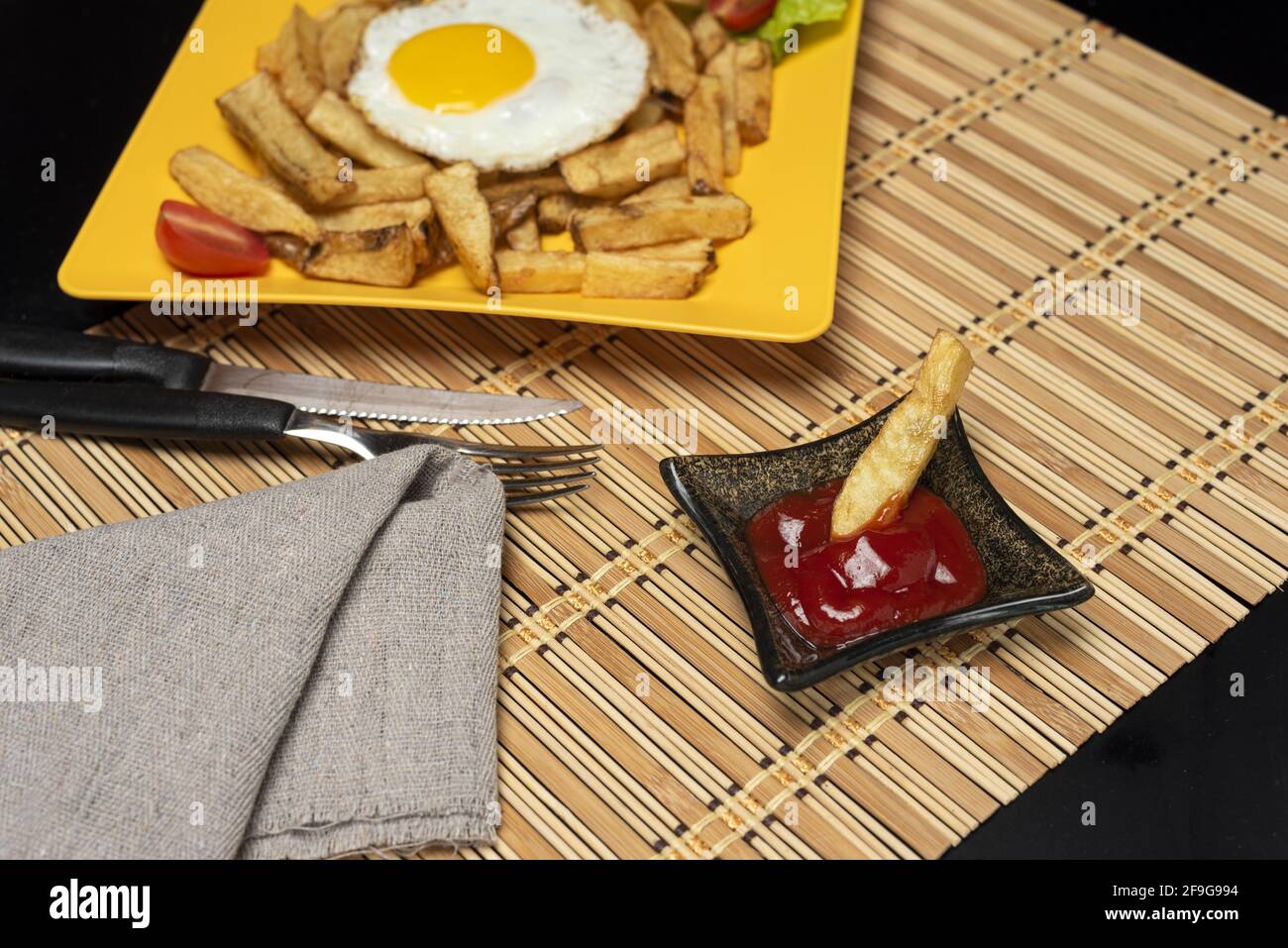 A high angle shot of fries and sunny side up egg on a yellow plate, with a single fry in the small bowl of ketchup Stock Photo