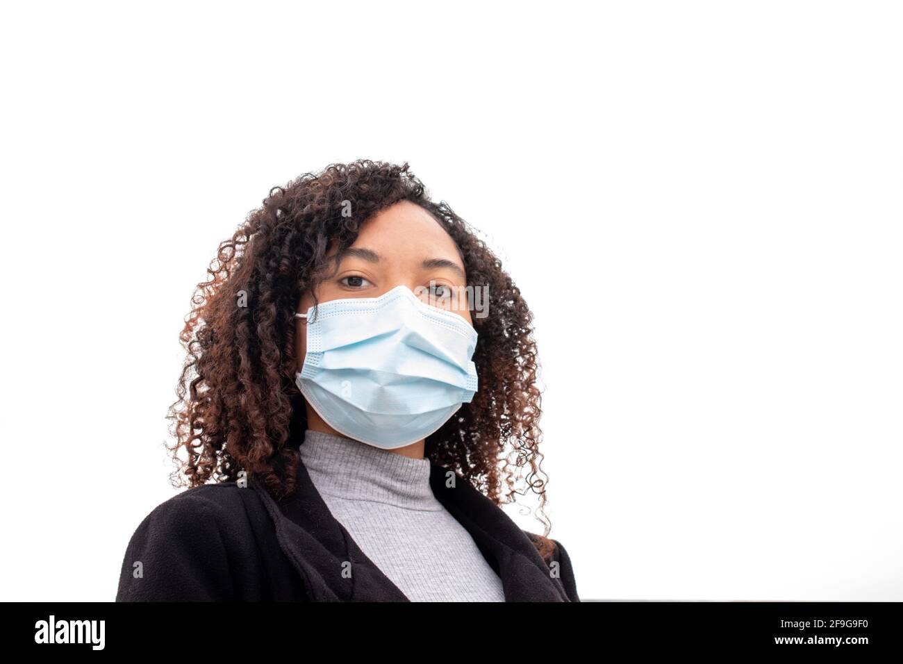 Beautiful dark-skinned young woman with curly hair wearing a protective mask against the COVID virus looks confidently at the camera. Stock Photo