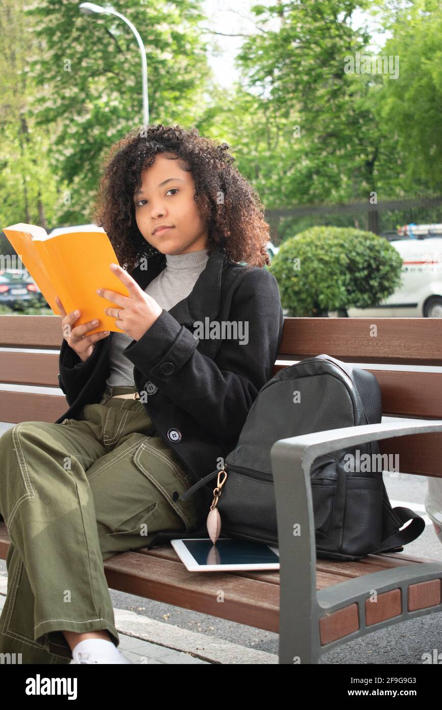 Young woman with curly hair and reading a book on a bench next to her are a backpack and a tablet. Stock Photo